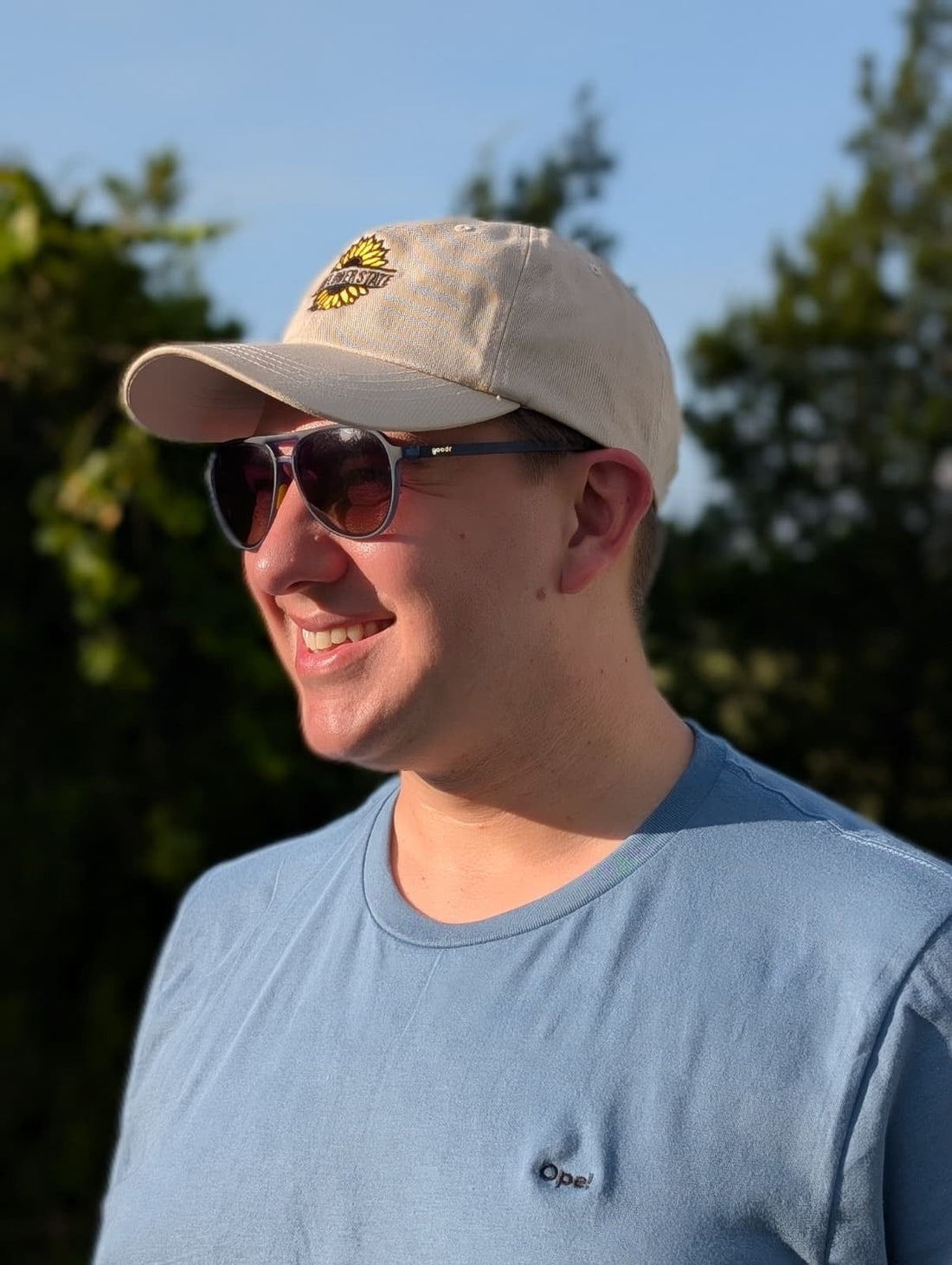 A person wearing sunglasses and a beige cap smiles warmly in a sunlit outdoor setting, surrounded by greenery and a clear blue sky.