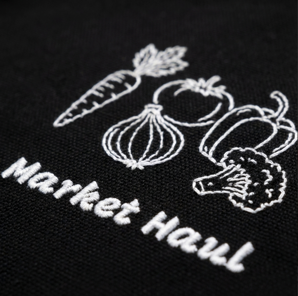 Detailed view of professional-grade embroidery on organic cotton twill, showing the texture of the thread and the clean line art of the farmstand vegetable design.