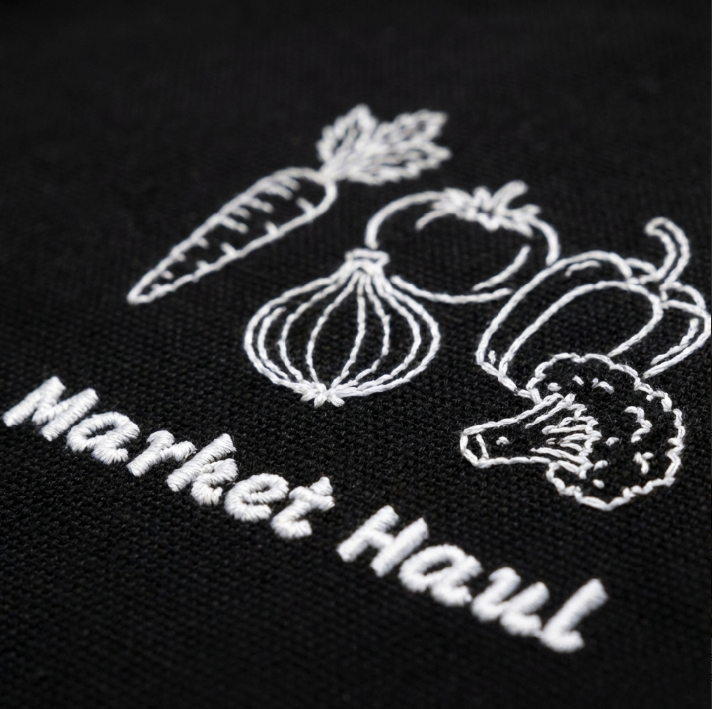 Detailed view of professional-grade embroidery on organic cotton twill, showing the texture of the thread and the clean line art of the farmstand vegetable design.